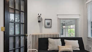 Una habitación con un sofá negro, una cesta de mimbre, un radiador y una ventana con persianas.
