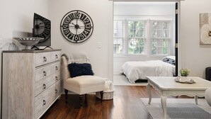 Un dormitorio con un aparador de madera, un reloj de pared redondo, una cama con ropa blanca y una ventana con persianas.