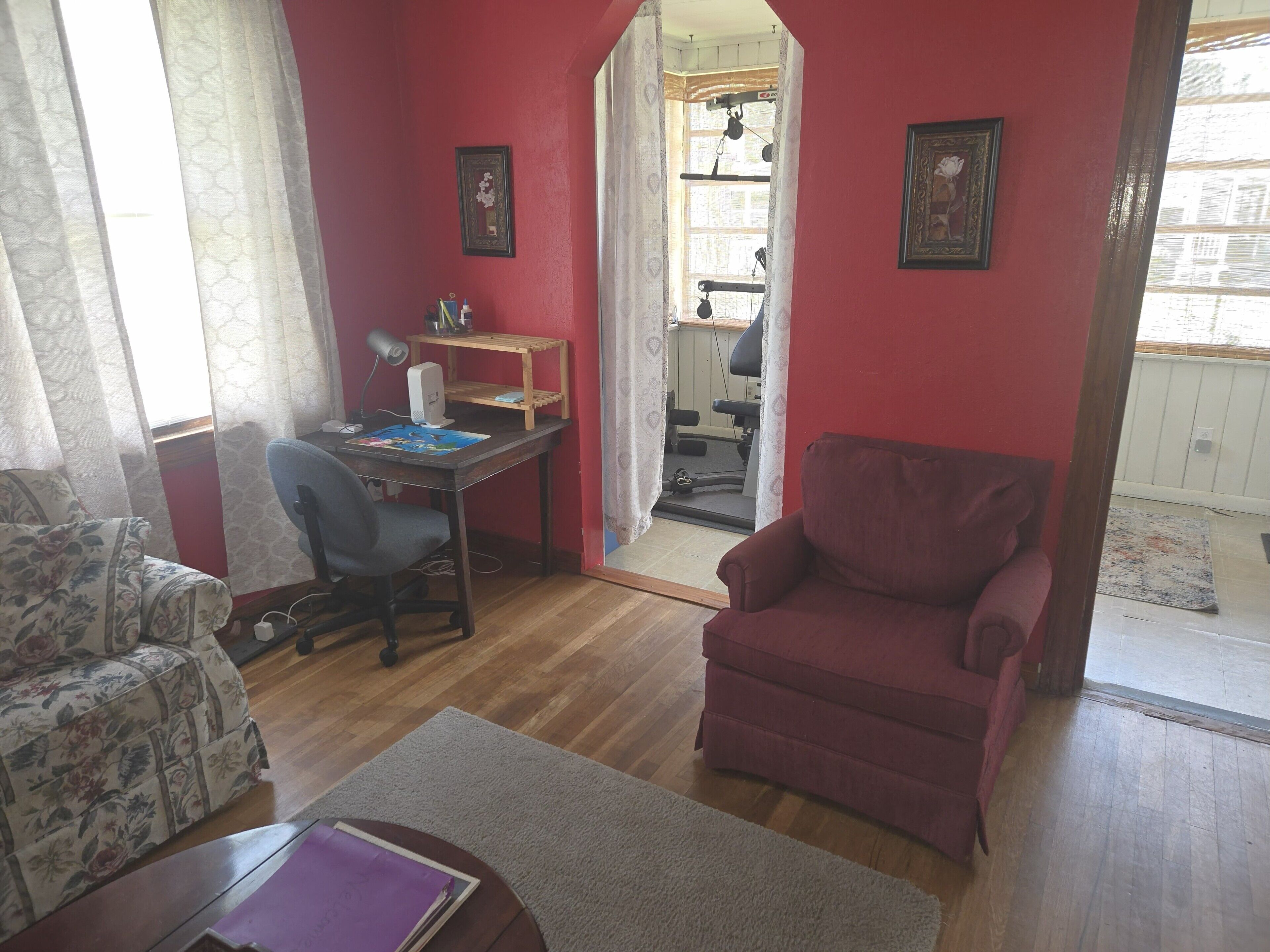 Una sala de estar con una pared roja, un escritorio de madera con una computadora, una silla y un sillón rojo. Hay una ventana con cortinas y una puerta que da a otra habitación.
