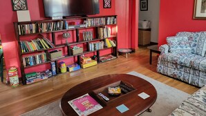Un salon avec un mur rouge, une bibliothèque remplie de livres et de jeux, un téléviseur à écran plat, un canapé à motifs floraux et une table basse avec des magazines.