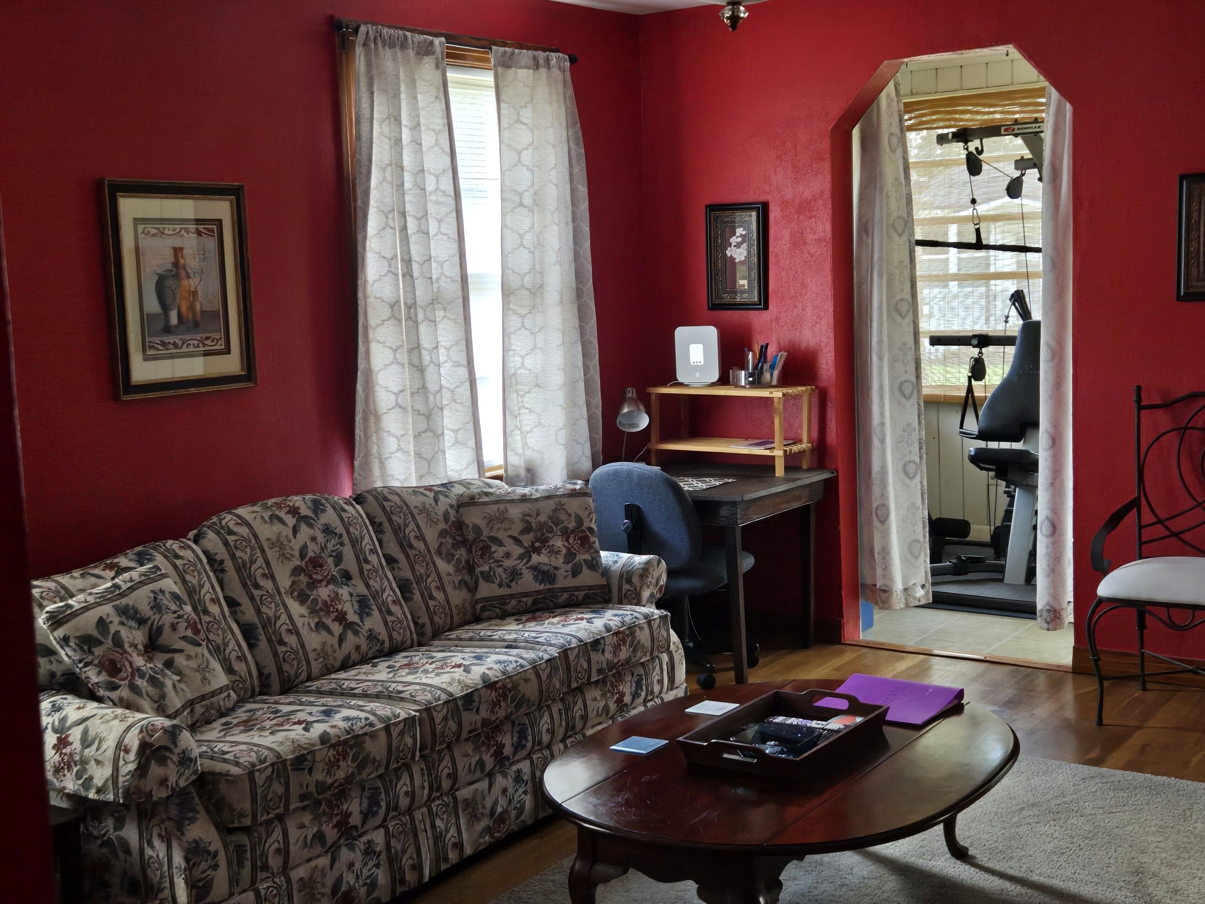 Un salon avec un canapé à motifs floraux, une table basse en bois, un bureau avec un ordinateur et, au fond, un tapis de course.