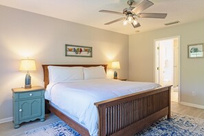 A bedroom with a wooden bed, two bedside tables with lamps, a ceiling fan, and a door leading to another room. - Oh Buoy! Bungalow- Family & Pet Friendly Beach House (Saint Augustine)