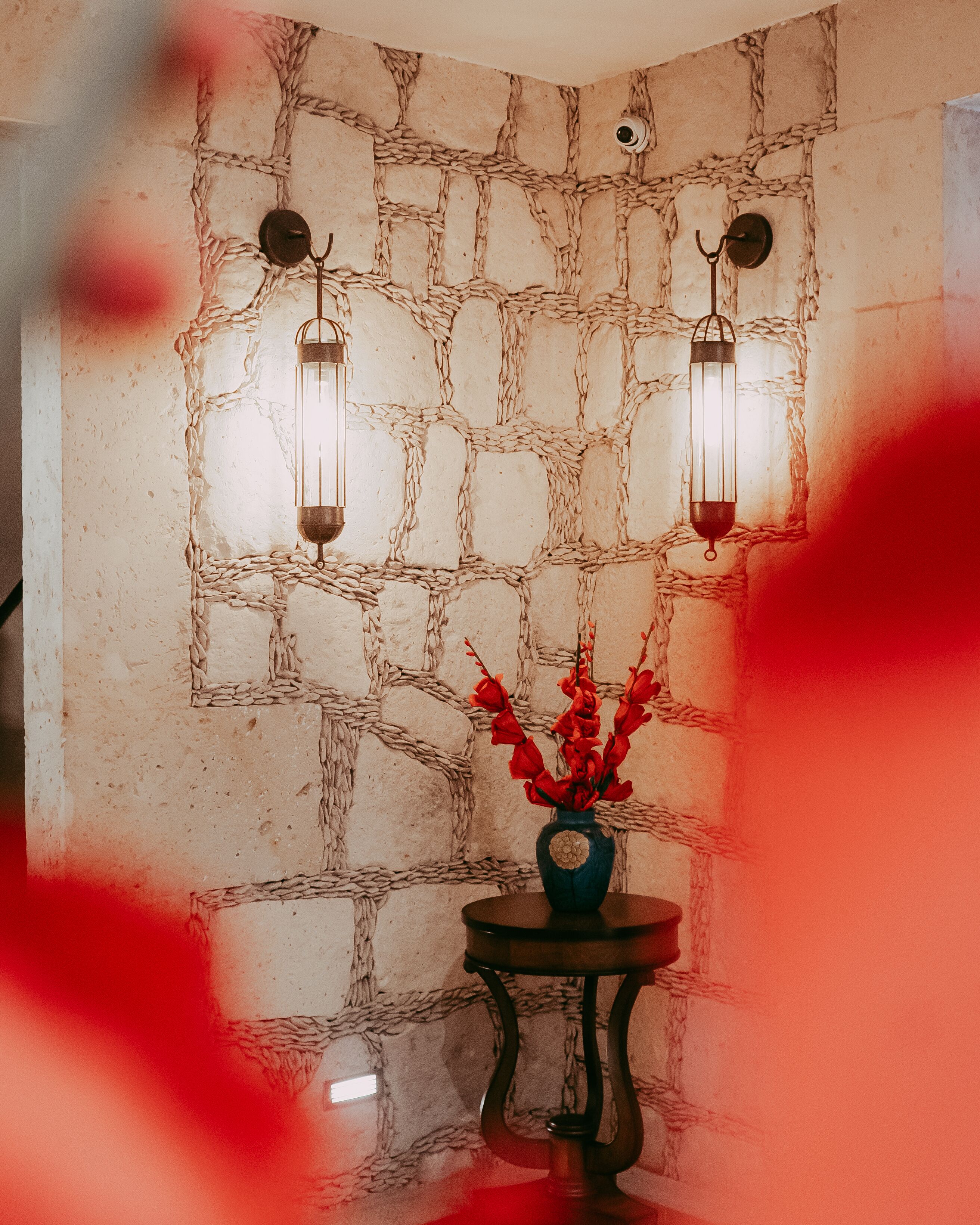 Un muro de piedra con dos faroles fijados a la pared y un jarrón con flores rojas sobre una mesita.