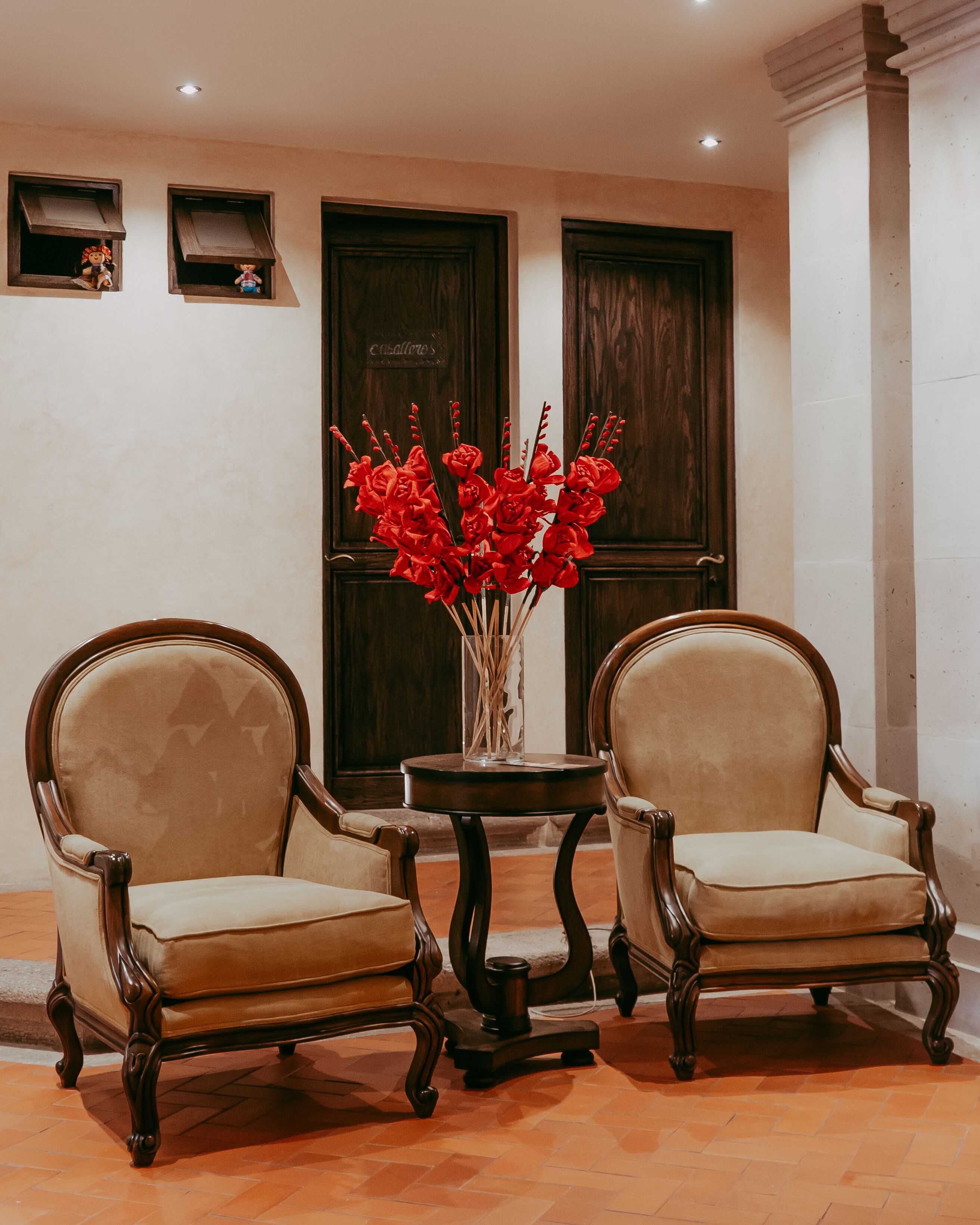 Deux fauteuils rembourrés à la structure en bois, et une petite table sur laquelle est posé un vase rempli de fleurs rouges, placée entre les deux fauteuils.
