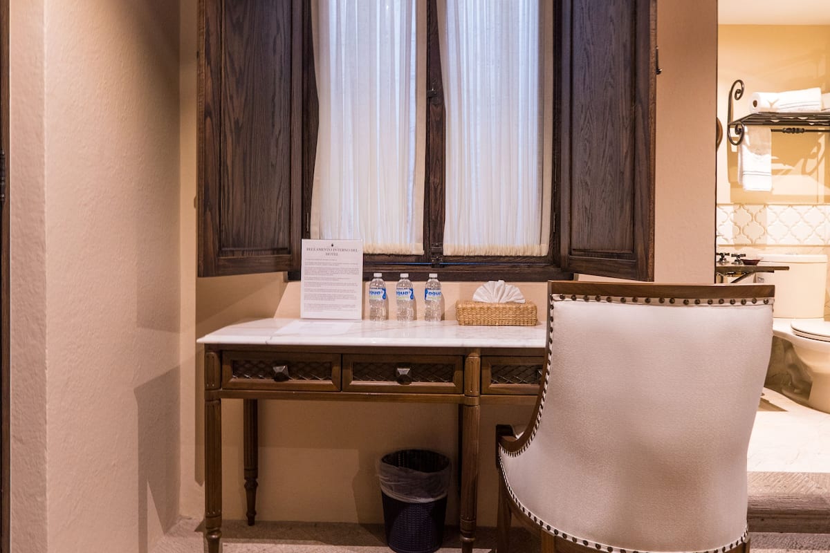 Een compacte, privé badkamer met een houten bureau, een stoel, een raam met gordijnen en een prullenbak.
