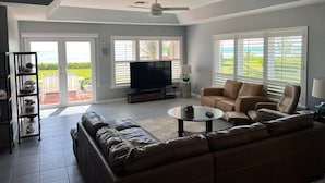 A living room with a large leather sofa, a television, and a coffee table. There are large windows with blinds, a ceiling fan, and a door leading to an outdoor area.