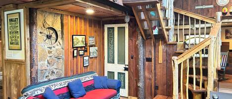 Un intérieur rustique avec un escalier en bois, un canapé agrémenté de coussins colorés et un mur décoré de tableaux encadrés et d’une horloge.