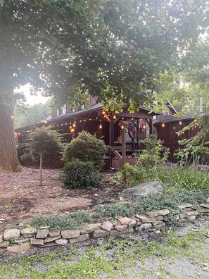 A rustic wooden cabin with a porch, surrounded by trees and a stone wall.