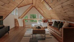 A spacious loft with a high ceiling, wooden beams, a cozy seating area with a sofa and a coffee table, and a balcony with a railing. - Firefly Cabin at Out of Bounds Retreats (Banner Elk)