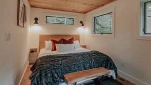 A bedroom with a wooden ceiling, a bed with a dark blanket, two bedside lamps, and a bench at the foot of the bed. - Firefly Cabin at Out of Bounds Retreats (Banner Elk)