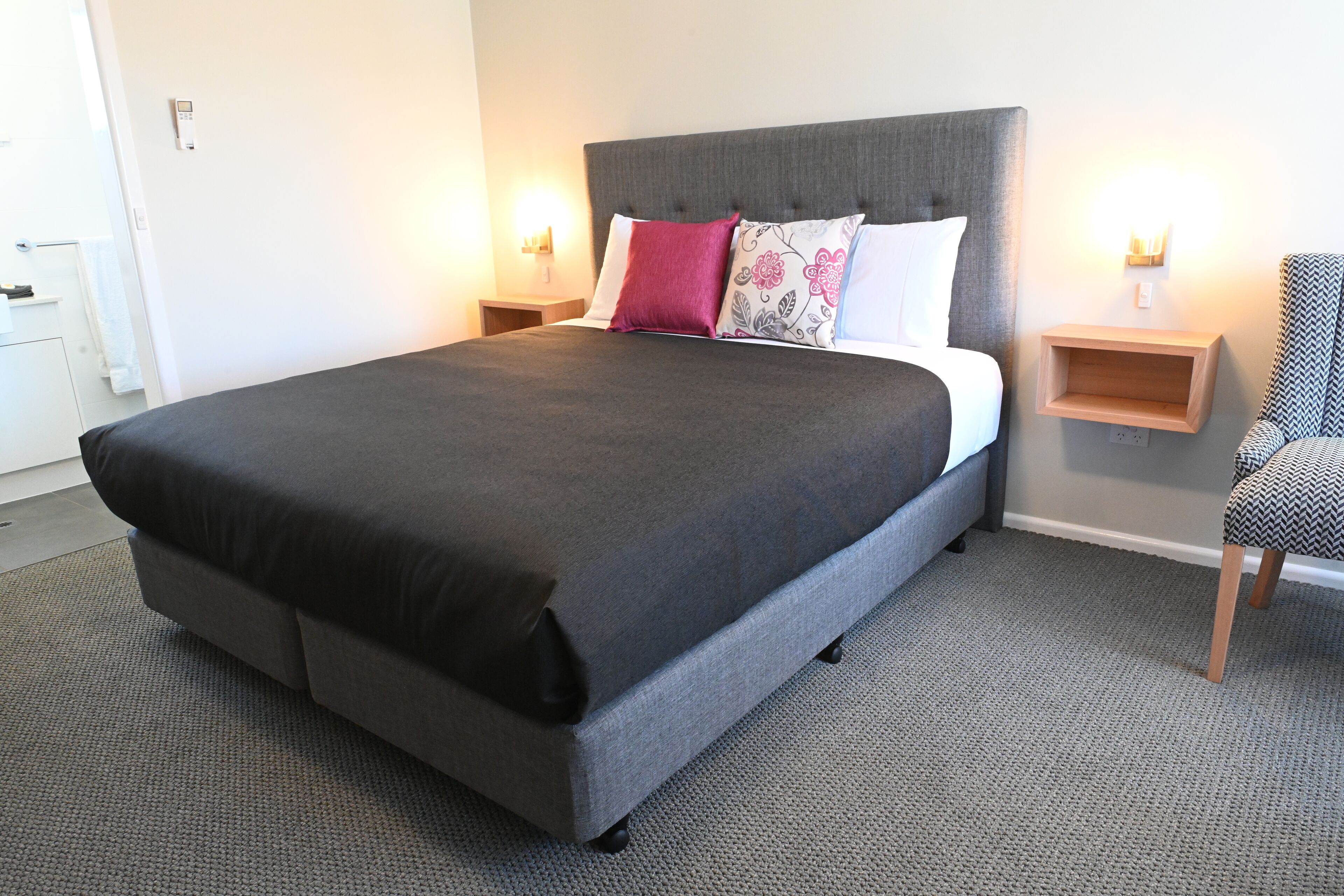 A modern hotel room with a large bed, grey upholstered headboard, and two bedside tables with lamps.