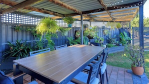 A covered patio area with a wooden table and chairs, surrounded by potted plants and a wooden fence.