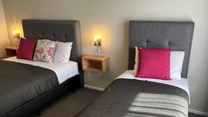 Two twin beds with grey headboards, a nightstand with a lamp, and a small wooden shelf.