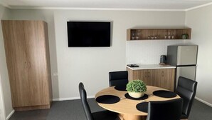 A compact living space with a round wooden dining table surrounded by black chairs, a flat-screen TV mounted on the wall, a kitchen area with a refrigerator, and a wooden wardrobe.