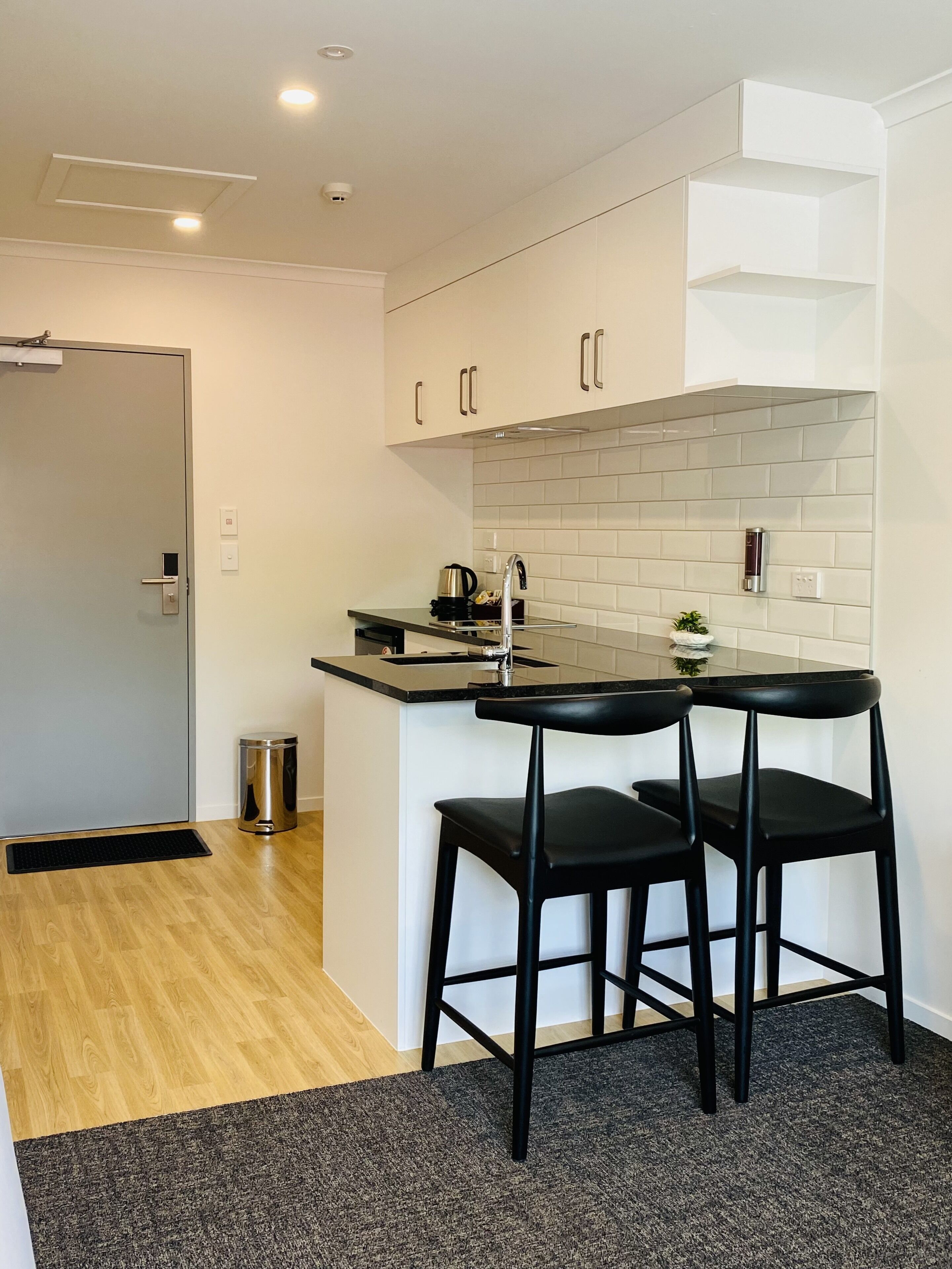 Una cocina compacta con encimera negra, dos taburetes negros y salpicadero blanco de azulejos.