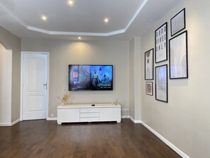 Uma sala de estar com uma TV de tela plana fixada na parede, um móvel para entretenimento branco embaixo e piso de madeira.
