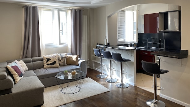 A modern living room with a grey sofa, a glass coffee table, and a kitchen area with a bar and three stools. - Bel Appartement à 12min de Paris (Colombes)