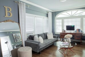 A living room with a grey sofa, a glass coffee table, a wooden desk, and a large window with blinds.