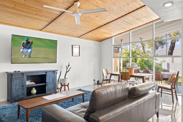 A modern living room with a large TV, leather sofa, wooden ceiling, and large windows.