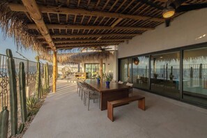 An outdoor patio with a wooden table and chairs, surrounded by cacti and a thatched roof structure.
