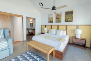 Ein Hotelzimmer mit Bett, einer Bank, einem Nachttisch, einem Deckenventilator und Kunstwerken an der Wand.