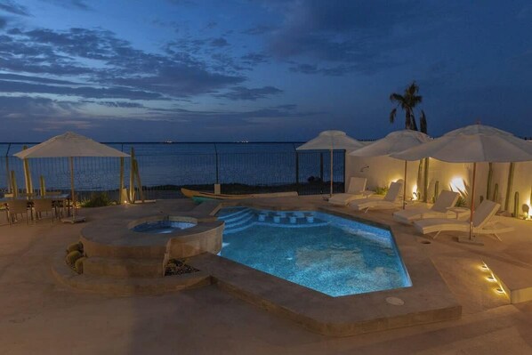 A pool area with lounge chairs and umbrellas, a hot tub, and a view of the sea at dusk.