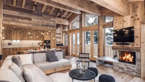 A spacious living room with a large sofa, a fireplace, a television, and a kitchen area with wooden walls and large windows.