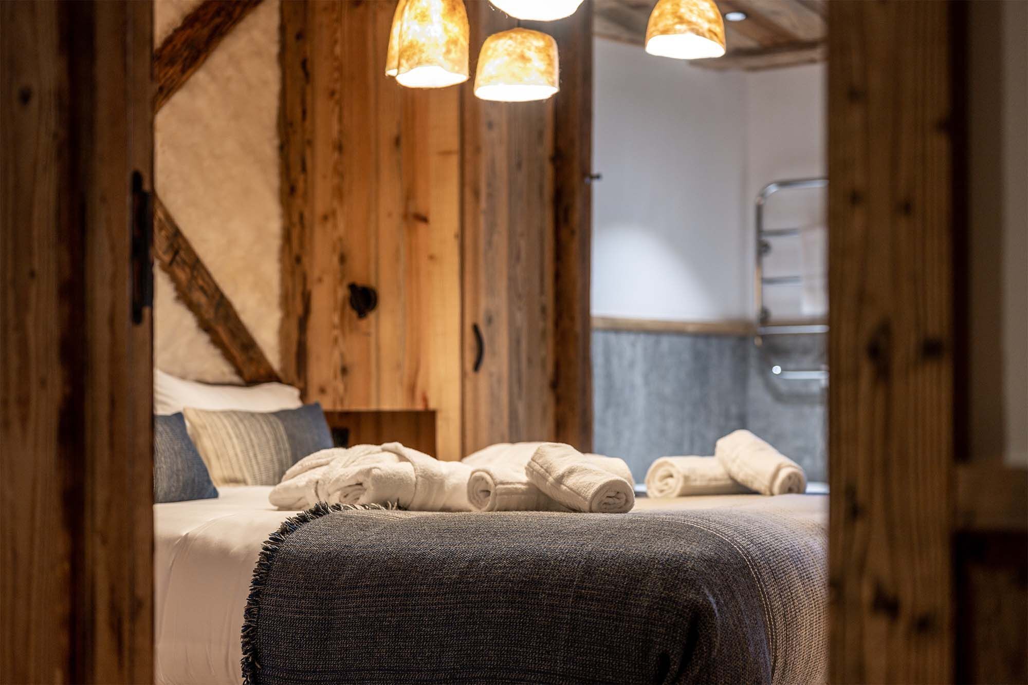 Une pièce chaleureuse avec des poutres apparentes, un lit agrémenté de serviettes roulées et un luminaire suspendu.