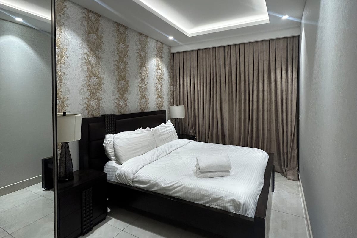 A modern bedroom with a large bed, a bedside table, a mirror, and a ceiling light.