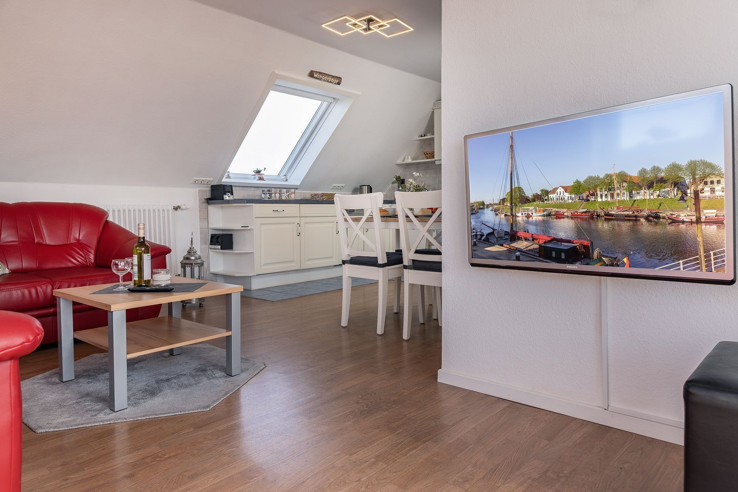 A modern living room with a red sofa, a wooden coffee table, and a dining area with white chairs. A large window provides a view of a canal with boats.
