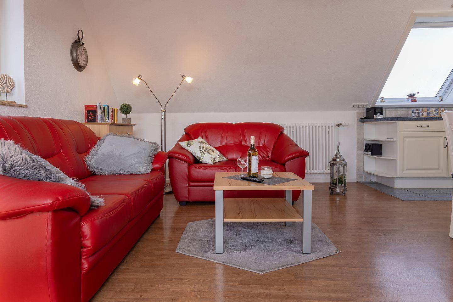 Een moderne woonkamer met een rode leren bank, een bijpassende fauteuil en een houten salontafel. Ook zichtbaar is een vloerlamp en een wandklok.