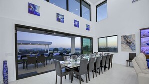 Un espace repas moderne avec une longue table dressée pour six personnes, de grandes fenêtres offrant une vue sur l’extérieur et des œuvres d’art accrochées aux murs.