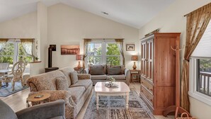 A spacious living room with a large sofa, a coffee table, and a wooden cabinet. There are two windows with curtains, a painting on the wall, and a rug on the floor. - Lovely Home- near fine dining and pool area (Bret Harte Cottage) (Angels Camp)