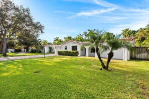 A residential property with a well-maintained lawn, a white house with a red-tiled roof, and a small garden area. - Retro 1950s Florida Home ~ Solar Heated Pool ~ Spa ~ Tiki Hut ~ Huge Backyard ~ Hammock ~ Two Bikes (Pompano Beach)