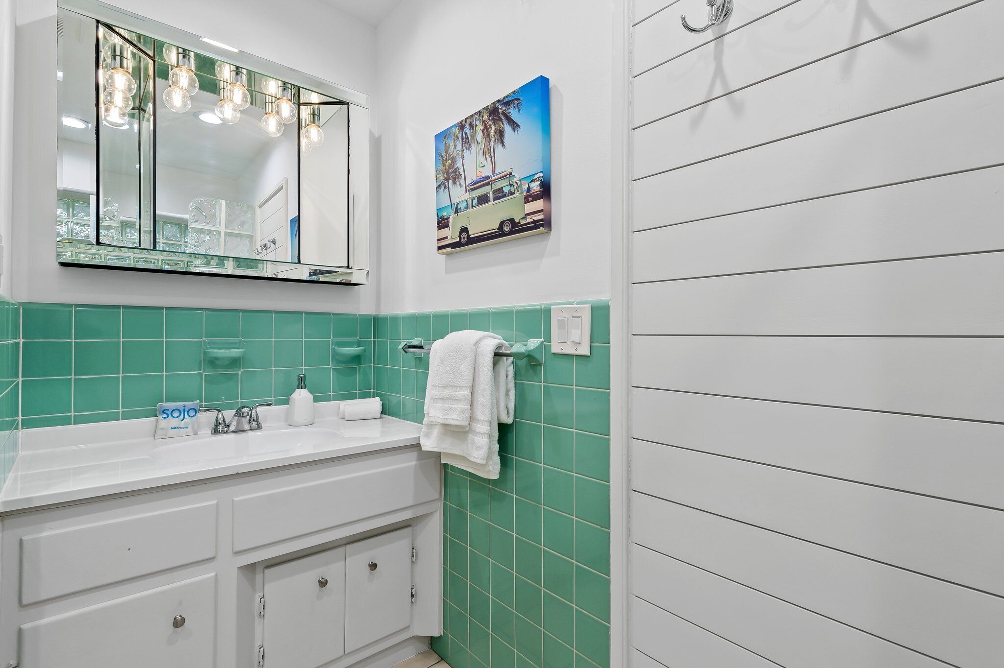 Un bagno con un lavandino bianco, piastrelle verdi, uno specchio illuminato, un portasciuga e un quadro raffigurante un furgone sulla parete.