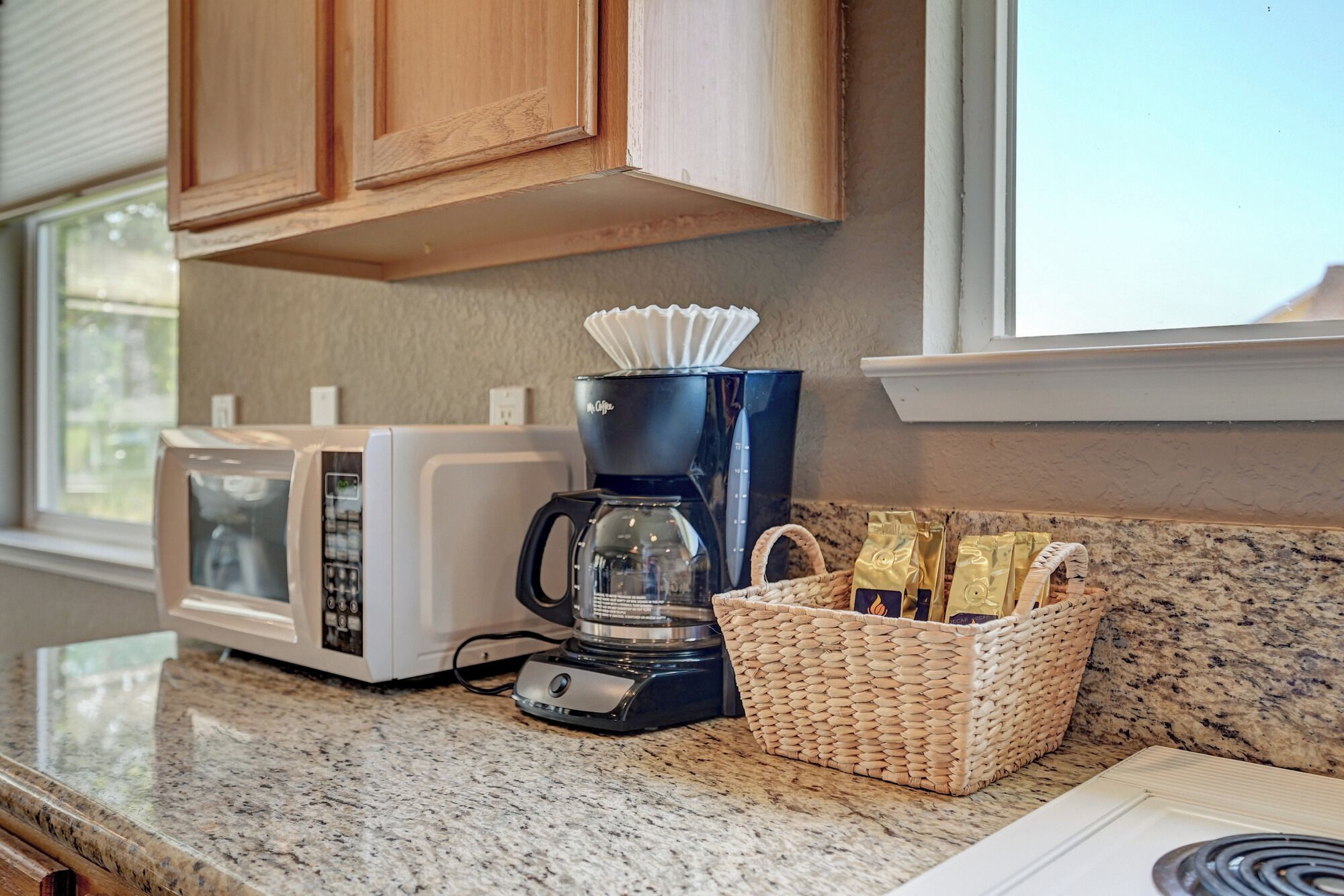 A kitchen with a coffee maker, microwave, and a basket of coffee packets on the counter.