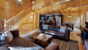 Una cabaña de madera espaciosa con una zona de estar central que incluye un sofá, una mesa de centro y una escalera que sube a un nivel superior.
