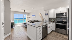 A modern kitchen with white cabinets, a granite countertop, stainless steel appliances, and a view to the ocean. - GULF FRONT BEDROOM @ CELADON BEACH RESORT ~FREE Beach Chair Service! (Panama City Beach)