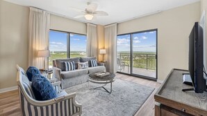 A spacious living room with a grey sofa, striped armchair, and a coffee table. Large windows offer a view of the outdoors.