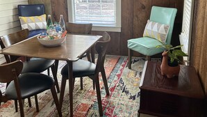 A dining area with a wooden table, chairs, a colorful rug, and a window with blinds. - Wensel Cottage in downtown Natchez Walking distance to most shops & restaurants. (Natchez)