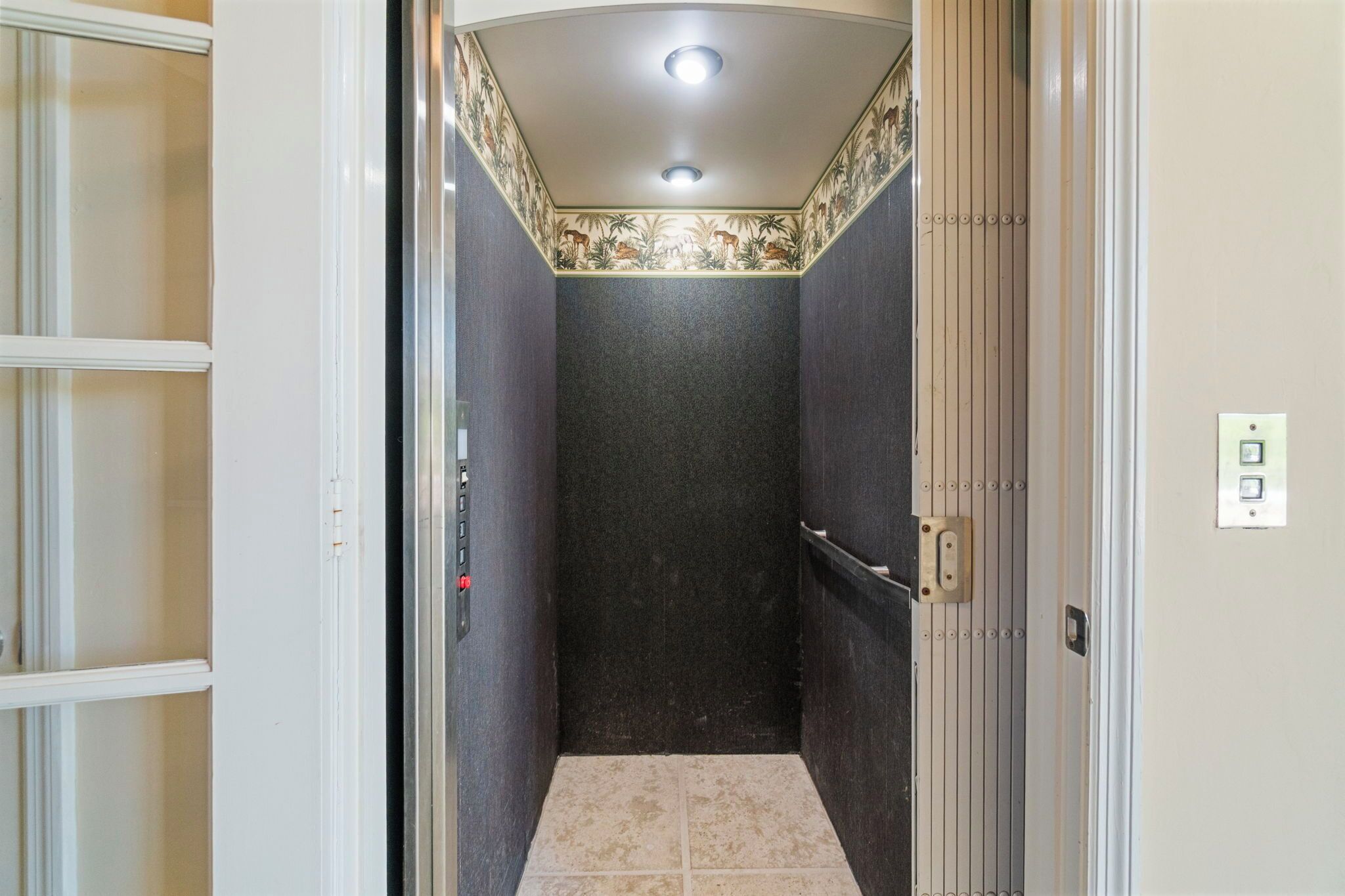 A narrow elevator shaft with a tiled floor, a ceiling light, and a door with a handle.