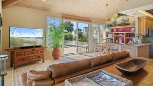 A spacious living room with a leather sofa, a television, and a kitchen area with wooden cabinets and a dining table.