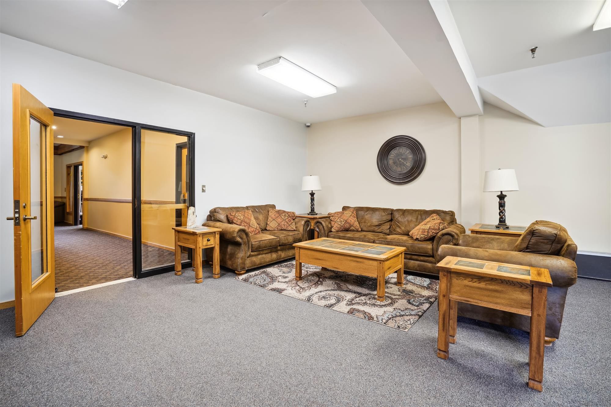 A spacious living room with a brown leather sofa set, two wooden side tables, a patterned rug, and a wall-mounted decorative plate.