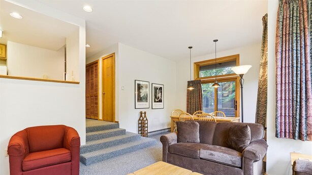 A living room with a red armchair, a brown sofa, and a wooden coffee table. There are stairs leading to a wooden door, and a window with curtains. - 2-bedroom 2-bath condo with pool, hot tub, and private sauna. (Killington)
