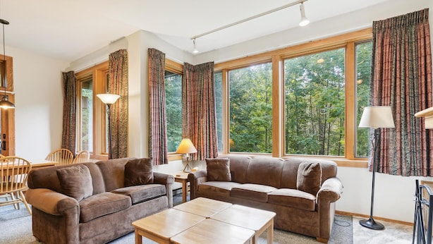 Ein gemütliches Wohnzimmer mit einer braunen Sofagarnitur, einem Holztisch und großen Fenstern mit Vorhängen.