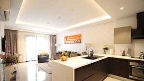 A modern kitchen with a central island, a dining area with a sofa and chairs, and a living room with a TV and curtains. - Mirage Residences - 2 Bedroom Unit (Accra)