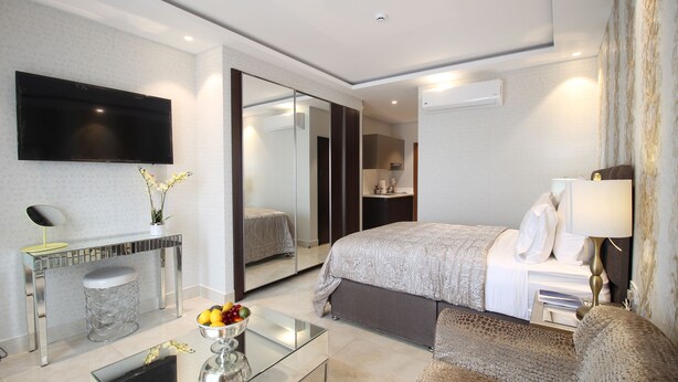 A modern hotel room with a large bed, a bedside table with a lamp, a glass coffee table, a mirror, and a TV mounted on the wall. - Mirage Residences - 2 Bedroom Unit (Accra)
