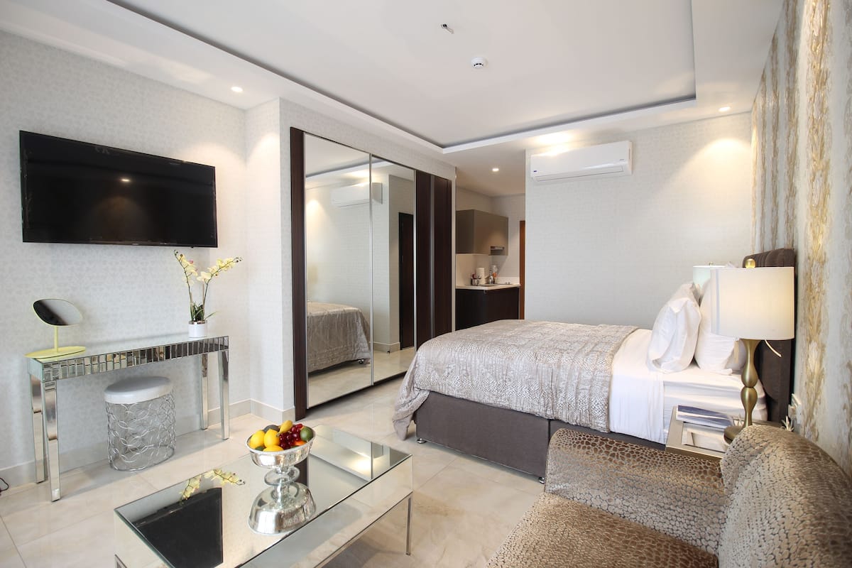 A modern hotel room with a large bed, a bedside table with a lamp, a glass coffee table, a mirror, and a TV mounted on the wall.