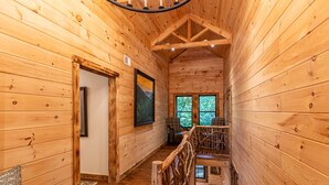 Interior de una cabaña de madera con escalera, paredes de madera, un cuadro enmarcado y ventana con vista a árboles.