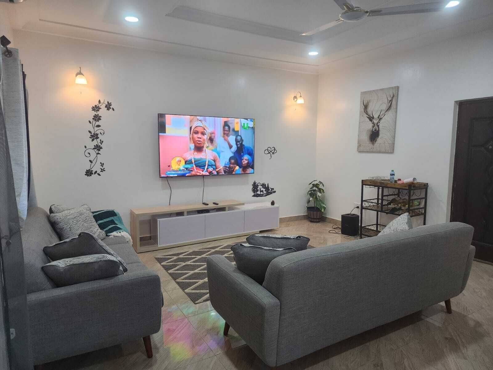 A living room with grey sofas, a flat-screen TV, a wooden cabinet, decorative wall art, and a ceiling fan.